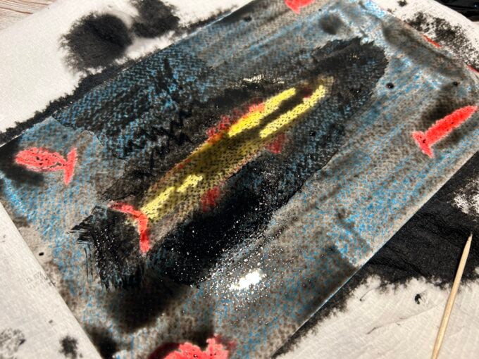 Painting over oil pastel fish with black paint for resist art technique